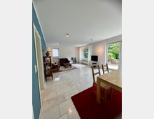 Großzügiger Wohn- und Essbereich mit hellen Fliesen, weißer Decke und Spotbeleuchtung, Sitzgruppe und Vitrine im hinteren Bereich sowie Esstisch auf rotem Teppich im Vordergrund. Große Fenster und eine Terrassentür lassen Blick ins Grüne; ein Fernseher steht an der rechten Wand.