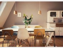 Esszimmer mit einem weißen Tisch, umgeben von verschiedenen Stühlen. Im Hintergrund stehen eine hellgraue Wand, ein weißer Schrank mit Holzelementen und zwei hängende Holzlampen; auf dem Tisch steht eine Vase mit grünen Pflanzen und Blüten.