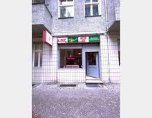 Eine kleine Ladenfront mit einer hell gefliesten Fassade in einem Wohngebäude. Über dem Schaufenster und der Eingangstür befinden sich Werbeschilder mit Zeitungsnamen, darunter "BZ", "Der Abend", "Hör zu" und "Berliner Morgenpost"; die Tür ist blau, und das Fenster bietet Einblick in einen Innenraum mit rotem Mobiliar.