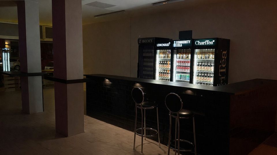 Dunkler Innenraum einer Bar oder Lounge mit langem Tresen und zwei hohen Barhockern. Hinter dem Tresen stehen beleuchtete Getränkekühlschränke, daneben sind Fenster und breite Säulen im Raum zu sehen.