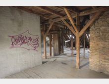 Alter Dachboden mit sichtbaren Holzbalken, Steinwänden und einem Holzboden. Auf einer grauen Wand ist ein violettes Graffiti zu sehen, im Hintergrund befinden sich Fenster und unordentliche Holzstrukturen.