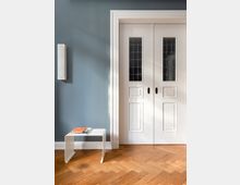 Weiße zweiflügelige Tür mit Glasfenstereinsätzen und dekorativem Rahmen vor einer hellblauen Wand; daneben ein minimalistischer, weißer Beistelltisch mit Büchern darauf sowie ein Parkettboden in Fischgrätmuster.