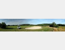 Golfplatz mit einem Sandbunker im Vordergrund, umgeben von gepflegten Rasenflächen und Bäumen; links steht ein weißes Golfmobil auf dem Grün. Der Himmel ist klar und blau.