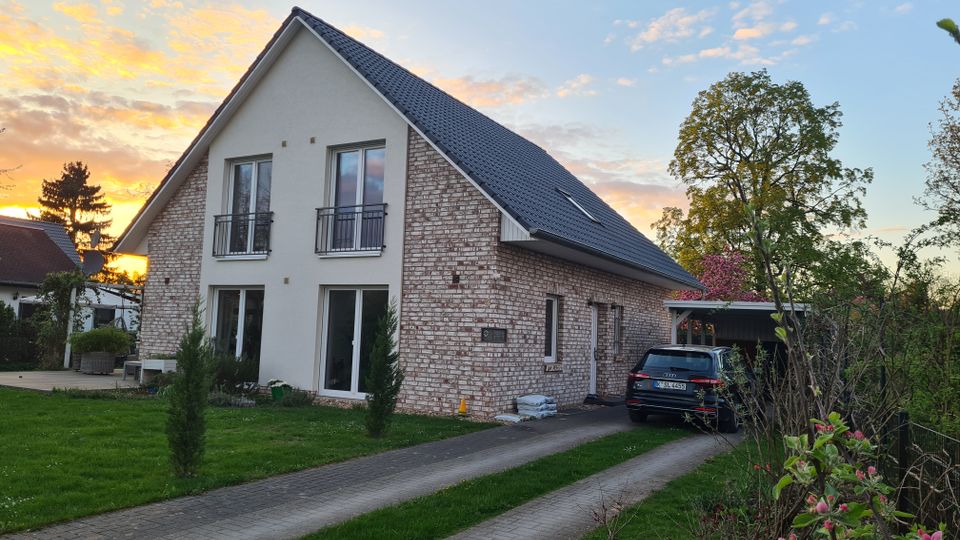 Ein modernes Einfamilienhaus mit weißer und rötlicher Klinkerfassade, Satteldach und bodentiefen Fenstern. Im Vordergrund ist ein gepflasterter Weg, der zu einer Garage mit geparktem Auto führt, umgeben von Rasen und Bäumen, während der Himmel im Hintergrund durch den Sonnenuntergang farbig erleuchtet ist.