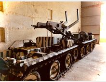 Ein militärisches Kettenfahrzeug mit Tarnbemalung steht in einer offenen Halle aus Beton. Die Halle ist spärlich beleuchtet, mit einer geöffneten Tür, durch die Tageslicht einfällt.