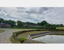 Grüne Parklandschaft mit gepflastertem Weg, einem kleinen rechteckigen Wasserbecken im Vordergrund und mehreren Backsteingebäuden im Hintergrund, umgeben von Bäumen und Hecken. Der Himmel ist bewölkt, und Lampen im traditionellen Stil säumen die Wege.