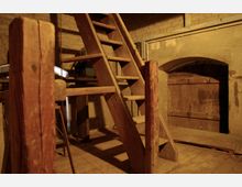 Holztreppe in einem alten, rustikalen Raum mit Steinwänden und Holzelementen. Im Hintergrund ist ein großer Holzbogen mit einer Holztür zu sehen.