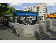 Eingang zur U-Bahn-Station "U-Bhf Richard-Wagner-Platz" mit blauem Schild, flankiert von einer Steinmauer und einer Rolltreppe, die nach unten führt. Im Hintergrund befinden sich Wohnhäuser, parkende Autos, ein Lieferwagen und ein Obststand.
