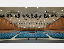 Großer moderner Veranstaltungssaal mit mehreren Reihen von blauen Sitzplätzen, einer erhöhten Bühne im Hintergrund und Holzvertäfelungen an den Wänden. An der Decke ist ein Beleuchtungssystem mit zahlreichen Scheinwerfern angebracht.