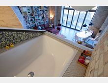 Ein modernes Loft mit Backsteinwänden und großen Fenstern, das eine offene Wohnfläche zeigt, einschließlich einer roten Couch, einer Bücherwand und einem Esstisch. Im Vordergrund ist eine geflieste Badewanne mit kleinen Regalen zu sehen.
