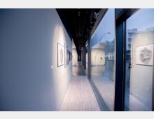 Ein moderner Galerieflur mit Holzboden und einer Reihe gerahmter Kunstwerke an der linken Wand, beleuchtet durch Spotlights. Rechts sind große Glasfenster, die Blick auf die Straße und umliegende Gebäude gewähren.