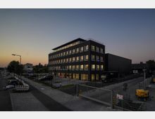 Moderne Büro- und Industriegebäude bei Abenddämmerung, mit eingeschaltetem Innenlicht. Das Gebäude hat eine dunkle Fassade, mehrere Stockwerke und ist von einem gepflasterten Hof mit geparkten Fahrzeugen und Bauzäunen umgeben.