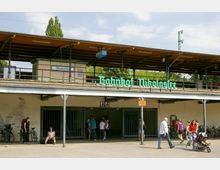 Bahnhof mit der Aufschrift „Bahnhof Nikolassee“, bestehend aus einem überdachten Bahnsteig mit Holz- und Metallkonstruktion und einer unteren Passage mit Eingängen. Im Vordergrund sind Menschen, Fahrräder und Kinderwagen zu sehen.