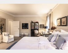 Helles Schlafzimmer mit einem großen Bett, weißer Bettwäsche und karierten Kissen, einem bequemen Sessel und einem Holztisch mit Dekorationen. Im Hintergrund sind ein Schrank mit Glastüren, Bilder an den Wänden und ein Fenster mit Gardinen sichtbar.