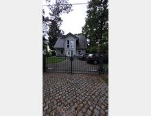 Einfamilienhaus mit grauer Fassade und Satteldach, das durch ein schmiedeeisernes Tor und eine gepflasterte Einfahrt zugänglich ist. Im Vordergrund ist ein mit Kopfsteinpflaster belegter Gehweg, während im Hintergrund Bäume das Grundstück umgeben.