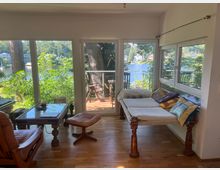 Wohnraum mit großen Fensterfronten, die Ausblick auf eine grüne, bewaldete Umgebung und einen See bieten. Im Vordergrund befinden sich Holzmöbel, darunter eine Liege mit bunten Kissen, ein Couchtisch und ein Ledersessel, sowie Zugang zu einem kleinen Balkon mit Sitzmöbeln.