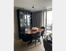 Moderner Essbereich mit ovalem Holztisch und gepolsterten Stühlen vor einem hohen, schwarzen Vitrinenschrank mit Glastüren und einer minimalistischen Pendelleuchte. Rechts öffnen bodentiefe Glastüren zum Balkon mit Vorhängen und Blick nach draußen.