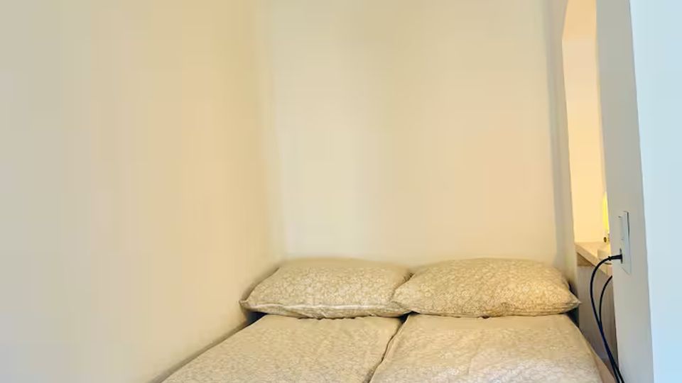 Kleines Schlafzimmer mit einem schmalen Doppelbett in einer Nische, weißen Wänden und hellem Holz-Bettgestell. An der Decke hängt eine geflochtene Rattanlampe; seitlich befindet sich ein arched Wanddurchbruch mit indirekter Beleuchtung.