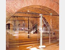 Gewölbter Innenraum aus roten Ziegelsteinen mit moderner Glaskonstruktion, die als Abtrennung dient. Der Boden besteht aus Holz, und eine Treppe führt zu einem weiteren Bereich im Hintergrund.