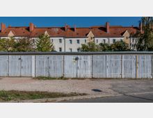 Reihe von blauen, verwitterten Garagentoren vor einem mehrstöckigen Wohngebäude mit roten Ziegeldächern und beige-weißer Fassade; im Hintergrund sind Bäume und Sträucher zu sehen, während der Vordergrund von einer Schotterfläche und einer Straße geprägt ist.