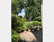 Ein gepflegter Garten mit einem gepflasterten Bereich im Vordergrund, auf dem mehrere Blumentöpfe mit grünen Pflanzen stehen. Im Hintergrund befinden sich ein Trampolin, eine Rasenfläche und große Bäume, die den Garten umgeben.