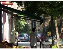 Eine städtische Straße mit dichtem Baumbestand, sichtbaren Autos und einem Backsteingebäude im Hintergrund. Im Vordergrund ein Gehweg mit einem Marktstand links, darüber hängt eine grüne Markise, und eine Person mit Kinderwagen läuft entlang.