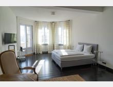 Helles, minimalistisches Schlafzimmer mit Doppelbett, dunklem Holzboden und großen Fenstern mit hellen Vorhängen in einer gebogenen Fensterfront. Links stehen ein Fernseher und ein kleiner Beistelltisch, daneben eine Glastür mit Fensterflügeln.