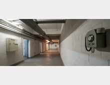 Ein langer, schlicht gehaltener Betonflur mit weiß gestrichenen Wänden und sichtbaren Lüftungsrohren an der Decke. An der rechten Wand hängt ein altmodisches Wählscheibentelefon, und links sind ein Sicherungskasten sowie offene Türen zu sehen.