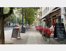 Straßenszene mit einem Gehweg, gesäumt von Bäumen und einem Café auf der rechten Seite mit Tischen im Außenbereich, angebotenem Mittagsmenü auf einer Tafel und Eimern voller bunter Blumen. Im Hintergrund sind Wohnhäuser und parkende Autos entlang der Straße zu sehen.