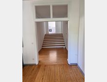 Innenraum mit Holzdielenboden, weiß gestrichenen Wänden und einer offenen Türöffnung, die zu einem kurzen Treppenaufgang mit fünf Stufen führt; im Hintergrund ist eine Küche mit weißen Möbeln sichtbar.