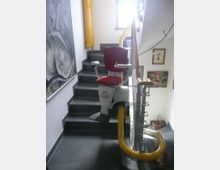 Treppenhaus mit schwarzen Fliesen und einem montierten Treppenlift mit rot gepolstertem Sitz auf einer gelben Schiene. An der Wand hängen Bilder, und es ist ein Fenster zu sehen, das Tageslicht hereinlässt.