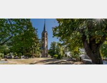 Kirche mit einem hohen Glockenturm aus Stein und einer spitzen Turmhaube, umgeben von großen Bäumen und einem kleinen Friedhof mit Grabsteinen. Im Vordergrund ist ein sonniger Grünbereich, während rechts eine Straße zu erkennen ist.