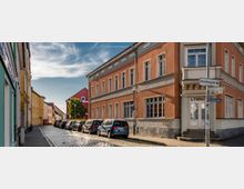 Eine gepflasterte Stadtstraße mit geparkten Autos und mehrstöckigen Gebäuden in warmen Farben, darunter ein orangefarbenes Eckgebäude mit großen Fenstern und klassischer Architektur. Straßenschilder und ein klarer Himmel ergänzen die Szene.