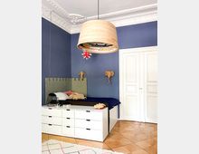 Bedroom interior with deep blue walls, ornate white ceiling molding, and a large cylindrical pendant light hanging over a bed with an upholstered headboard. A low white drawer unit sits at the foot of the bed, with white paneled double doors and a herringbone wood floor completing the room.