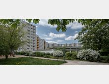 Wohngebiet mit mehreren Plattenbauten im Hintergrund, umgeben von Bäumen, blühenden Sträuchern und einem kleinen Parkbereich mit Wegen und einem Spielplatz. Der Vordergrund zeigt Rasenflächen und blühende Kastanien.