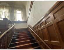 Holzgetäfeltes Treppenhaus mit einer hölzernen Balustrade und einem roten Teppichläufer auf den Stufen. Im Hintergrund befinden sich hohe Fenster mit halbtransparenten Vorhängen, die weiches Licht hereinlassen.