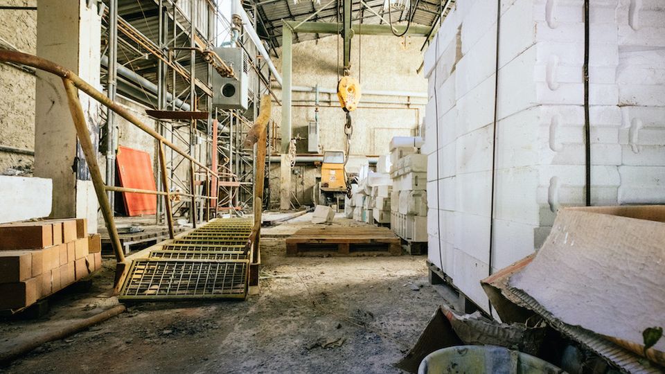 Industrielle Lagerhalle mit Metallgerüsten, gestapelten weißen Blöcken und Baumaterialien wie Ziegeln und Plastikeimern; im Hintergrund sind ein Deckenkran und Stahlträger sichtbar.