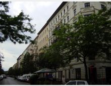 Straßenansicht mit klassischen Wohngebäuden aus der Gründerzeit, verziert mit Stuckfassaden. Am Straßenrand stehen geparkte Autos, und Bäume säumen den Gehweg; im Vordergrund befindet sich ein kleiner Kiosk mit Sonnenschirm.