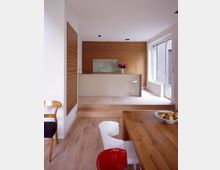 Moderne Küche mit Holzdetails, bestehend aus einer erhöhten Küchenzeile mit einer glatten, weißen Arbeitsfläche und einer Rückwand aus horizontalen Holzpaneelen. Der Raum hat helle Holzböden, ein großes Fenster, das viel Tageslicht hineinlässt, und eine angrenzende Essbereich mit einem Holztisch und farbigen Stühlen.