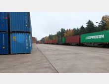 Großer Güterumschlagsplatz mit aufeinander gestapelten blauen Containern links und einem Güterzug mit roten und grünen Containern auf der rechten Seite, umgeben von herbstlich gefärbten Bäumen im Hintergrund.