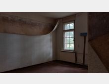 Ein leerer Raum mit abgeblätterter Tapete an den Wänden, einem Holzfenster mit Blick auf eine grüne Umgebung und einem alten Heizkörper darunter. Rechts an der Wand hängt ein Schild mit der Aufschrift "Telefunken Sendertechnik".