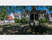 Denkmal auf einem kleinen Platz mit Stufenfundament, umgeben von Bäumen und Grünflächen. Im Hintergrund sind Wohnhäuser mit roten und grauen Dächern sowie eine gepflasterte Straße erkennbar.