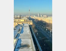 Blick über eine Straße in Berlin mit dem Berliner Fernsehturm im Hintergrund. Links im Vordergrund ein Flachdach mit leichtem Schnee, rechts eine mehrspurige Straße, gesäumt von Bäumen und historischen Gebäuden.
