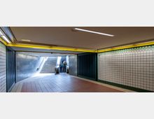 Unterführung mit gefliesten Wänden in weiß und grünem Muster, gelb beleuchteter Decke und Fliesenboden in Rot- und Beigetönen. Im Hintergrund führen Treppen und Rolltreppen nach oben, und Personen stehen im Bereich des Aufgangs.