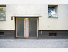 Eingangsbereich eines Wohnhauses mit doppelter Glastür und Hausnummer "65" über der Tür. Die Fassade ist hellgelb mit braunen Fliesen im unteren Bereich, und neben der Tür befinden sich Klingelschilder sowie ein Fenster auf jeder Seite.