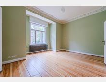 Leerer Wohnraum mit hellgrünen Wänden, breiten Holzdielen und einer hohen Decke mit Stuckverzierung. An der Stirnwand befindet sich ein großes Fenster über einem dunklen Heizkörper, daneben eine weiße Tür.