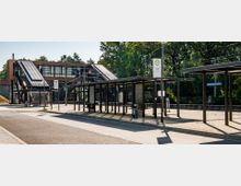 Bushaltestelle und Bahnhof in einer modernen Umgebung mit Überdachungen aus Metall, einer großen Glasfassade und Treppenaufgängen, die zu einem erhöhten Bahnsteig führen. Im Hintergrund sind Bäume und ein bewaldetes Gebiet sichtbar.