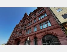 Historische Fassade eines mehrstöckigen Gebäudes aus rotem Sandstein mit dekorativen Verzierungen, großen Fenstern und einem stufenartigen Giebeldach; im Vordergrund eine Laterne. Der Himmel ist klar und blau.