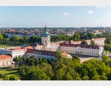 Luftaufnahme des Schlosses Charlottenburg in Berlin, mit seiner zentralen, mit einer grünen Kuppel gekrönten Fassade und weitläufigen, beige-gelben Gebäudeflügeln. Umgeben von Bäumen, Gärten und im Hintergrund eine städtische Skyline mit Gebäuden.
