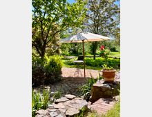 Garten mit gepflastertem Weg, schattiert durch Bäume und einen großen weißen Sonnenschirm. Im Hintergrund stehen ein Tisch und Stühle, umgeben von Rasenflächen, Blumenbeeten und einem Pflanzentopf aus Terrakotta.